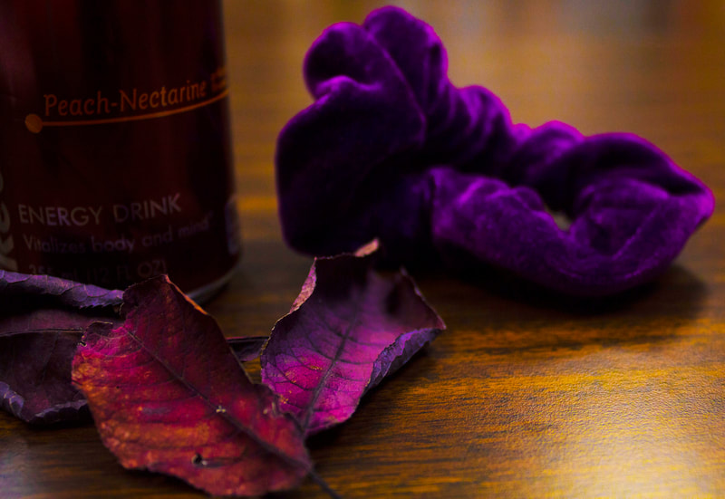 Curling purple leaves in front of a peach-nectarine redbull can and purple scrunchie